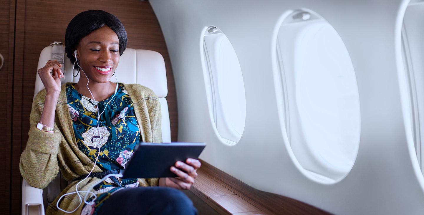 穿着印花衬衫的女人戴着连接平板电脑的耳机, 在私人飞机上享受机上娱乐