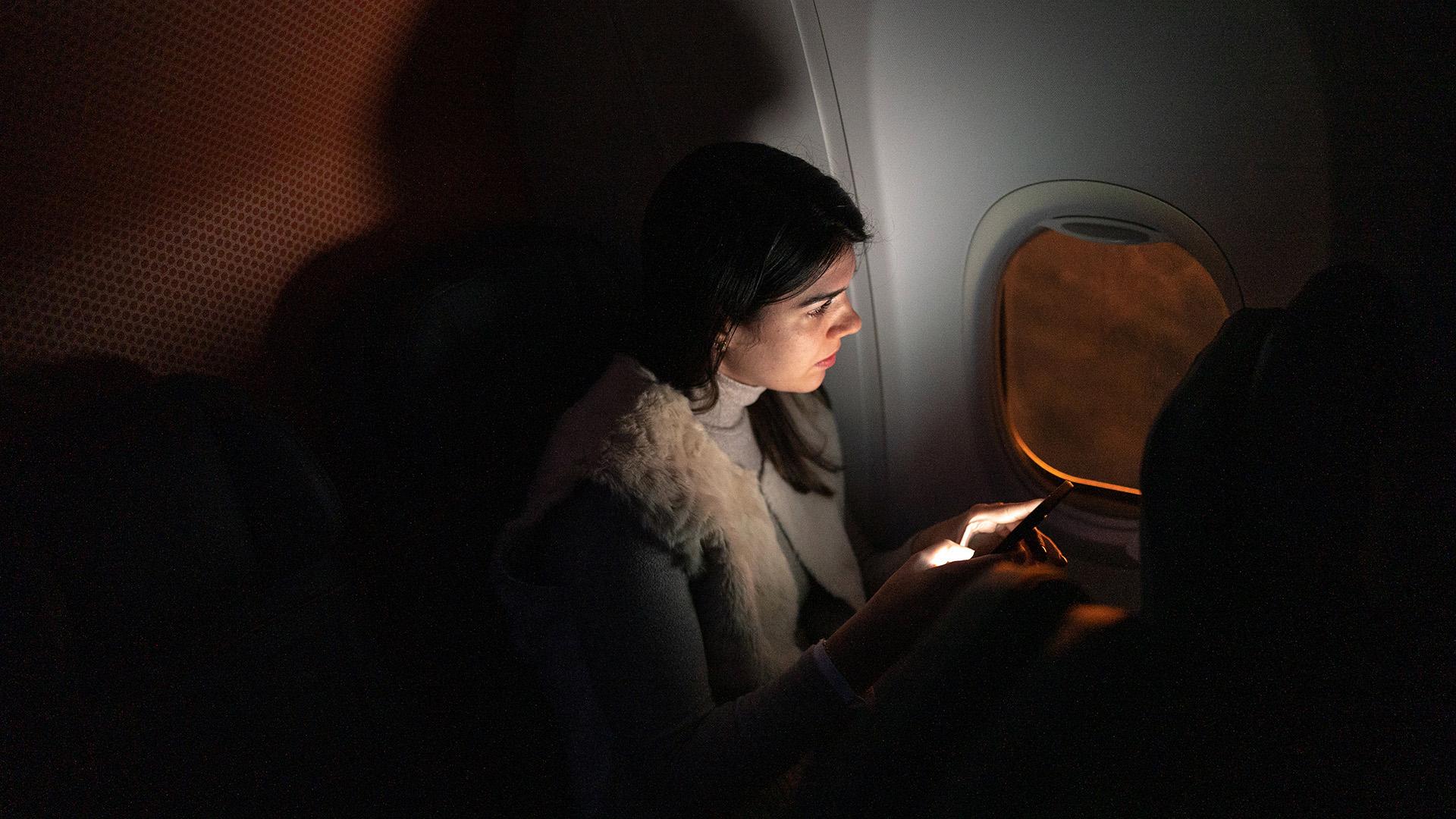 A passenger with long hair sits next to an airplane window in a dark cabin, looking out at a sunset, with the light from their phone illuminating their face.